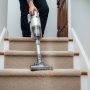 A lightweight cordless handheld vacuum being used to clean the carpet on a narrow residential staircase with beige low-pile carpet.