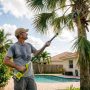 Homeowner using a cordless pole saw to trim tall palm tree fronds in a sunny Florida backyard
