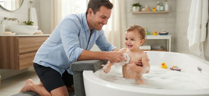 Father kneeling on a padded kneeler while bathing a toddler in a tub.